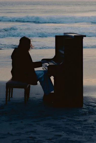 Lady playing piano on a beach with the sea beside her.